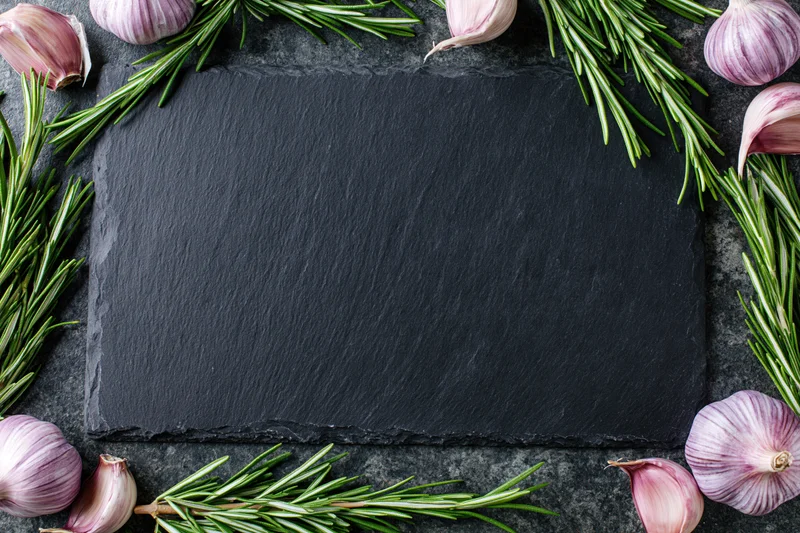 Black slate cutting board framed by garlic and rosemary sprigs on dark stone surface