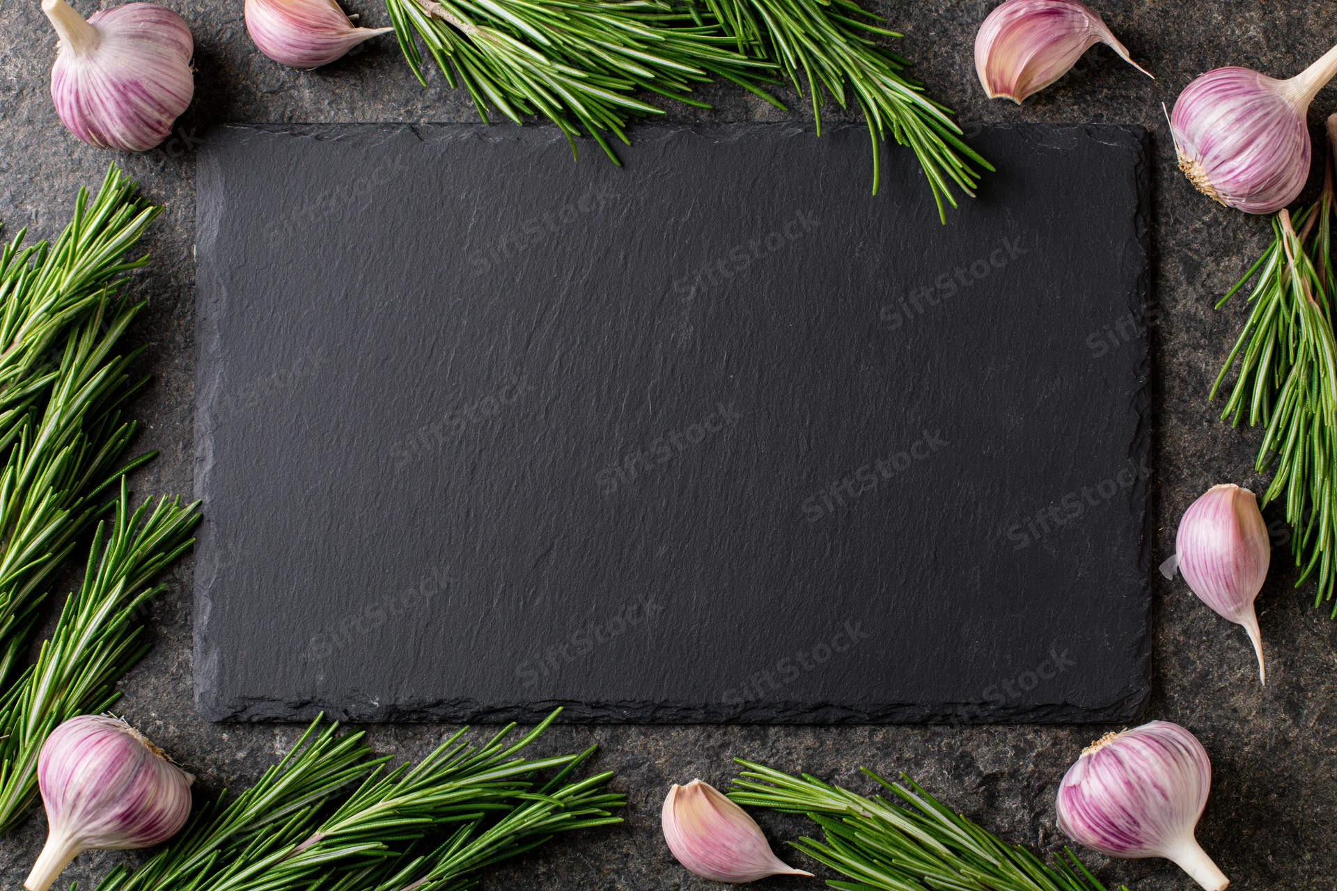 Black slate cutting board centered on dark stone countertop surrounded by garlic bulbs and rosemary sprigs in flat lay arrangement
