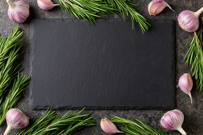 Black slate cutting board centered on dark stone countertop surrounded by garlic bulbs and rosemary sprigs in flat lay arrangement