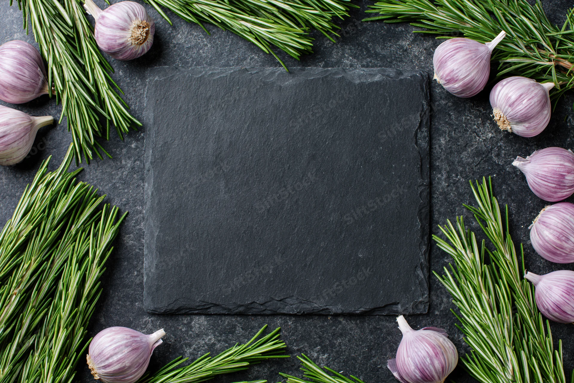 Black slate cutting board centered on dark stone countertop surrounded by garlic and rosemary sprigs in flat lay arrangement