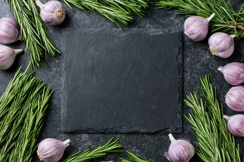 Black slate cutting board centered on dark stone countertop surrounded by garlic and rosemary sprigs in flat lay arrangement