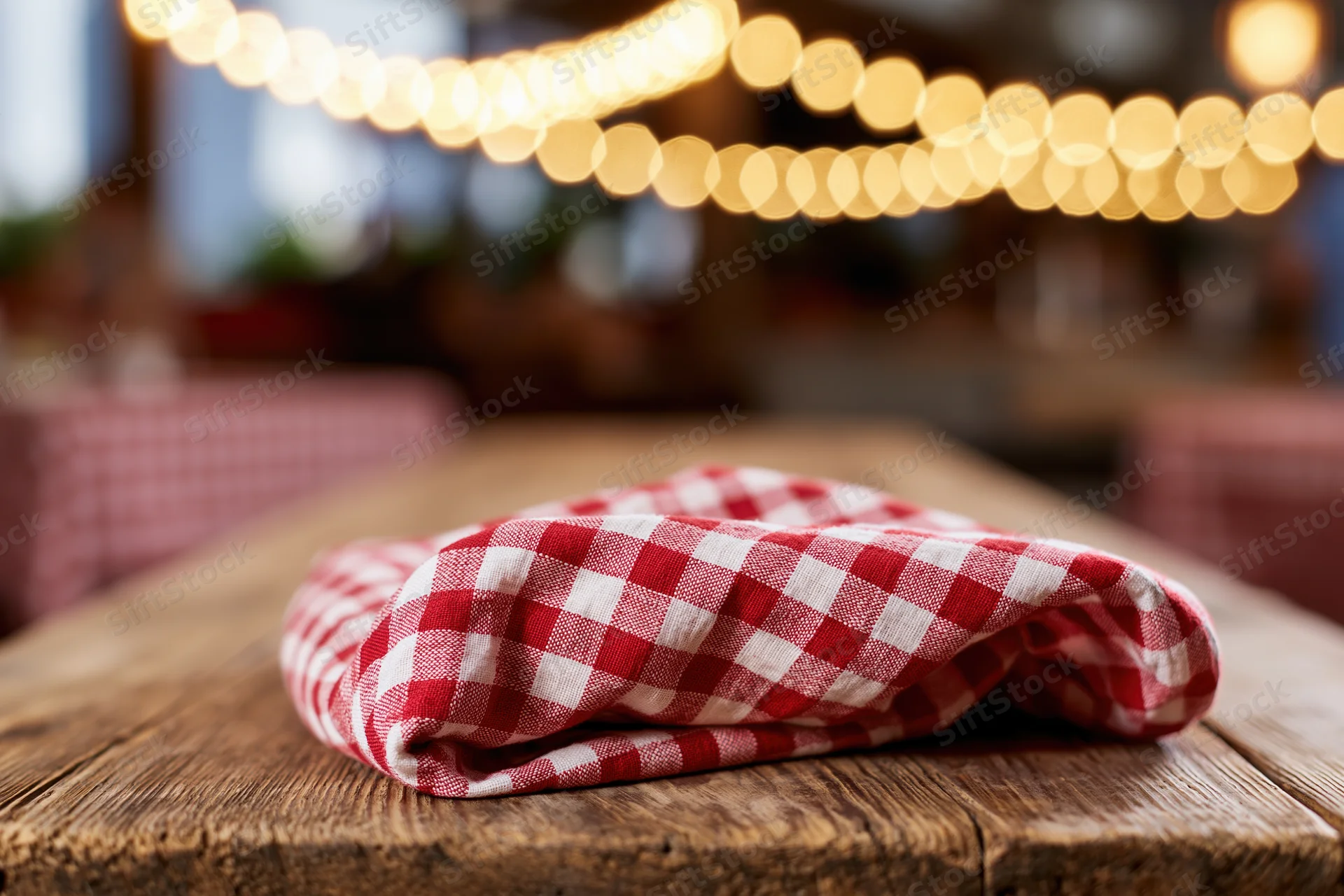 Folded red and white checkered napkin on rustic wooden table with string lights in background