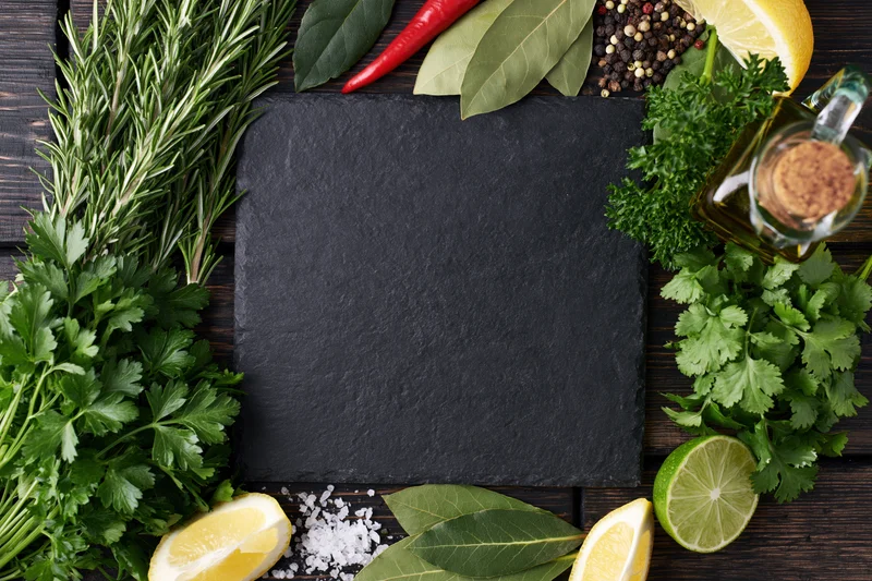 Black slate board centered on dark wooden table surrounded by fresh herbs, citrus, spices and olive oil in flat lay arrangement