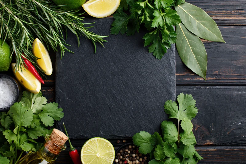 Black slate board surrounded by fresh herbs and spices on dark wooden surface