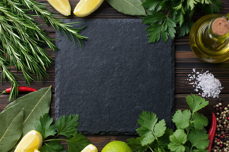 Black slate board centered on dark wooden table surrounded by fresh herbs, citrus, spices, and oil with copyspace