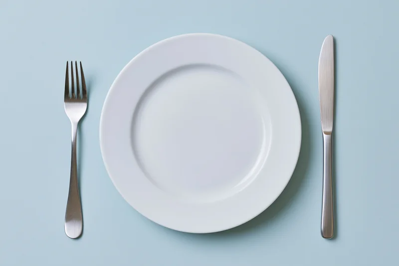 Empty white ceramic dinner plate with fork and knife arranged on pale blue surface in flat lay overhead view