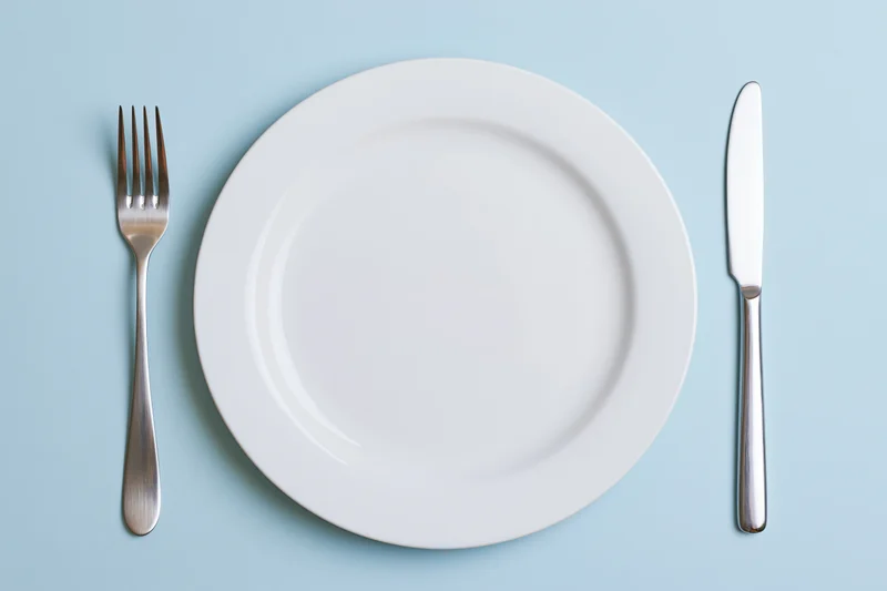 Empty white ceramic plate with fork and knife arranged on pale blue surface in flat lay view