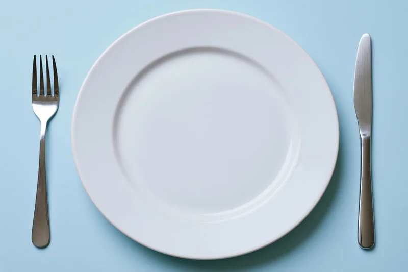 Empty white ceramic dinner plate with fork and knife on pale blue surface