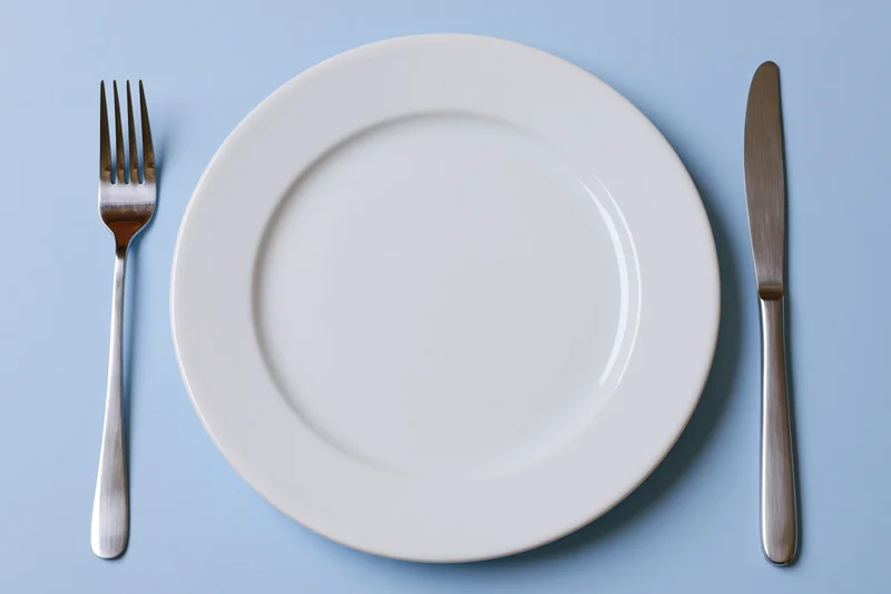 Empty white ceramic dinner plate with fork and knife on pale blue surface