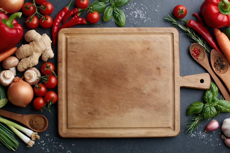 Wooden cutting board surrounded by fresh vegetables and herbs on dark slate surface