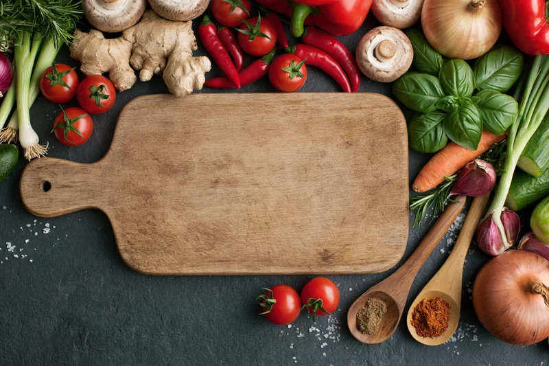 Wooden cutting board surrounded by fresh vegetables and herbs on dark slate surface