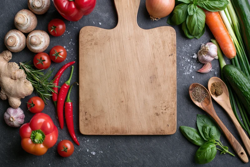 Wooden cutting board centered on dark slate surface surrounded by fresh vegetables and herbs for meal preparation