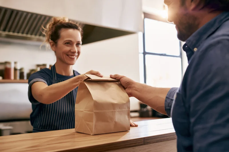 Woman in striped apron handing paper bag to customer at cafe counter