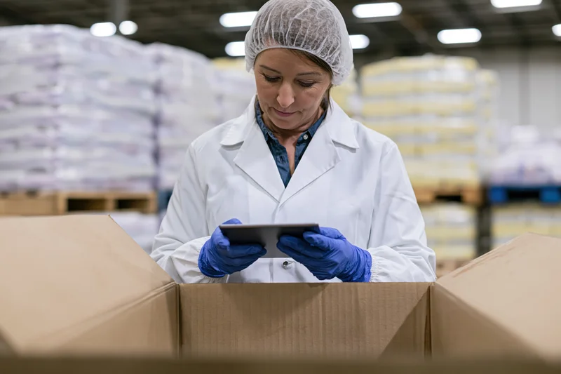 Woman in lab coat and hairnet inspecting a box with tablet in a food factory warehouse