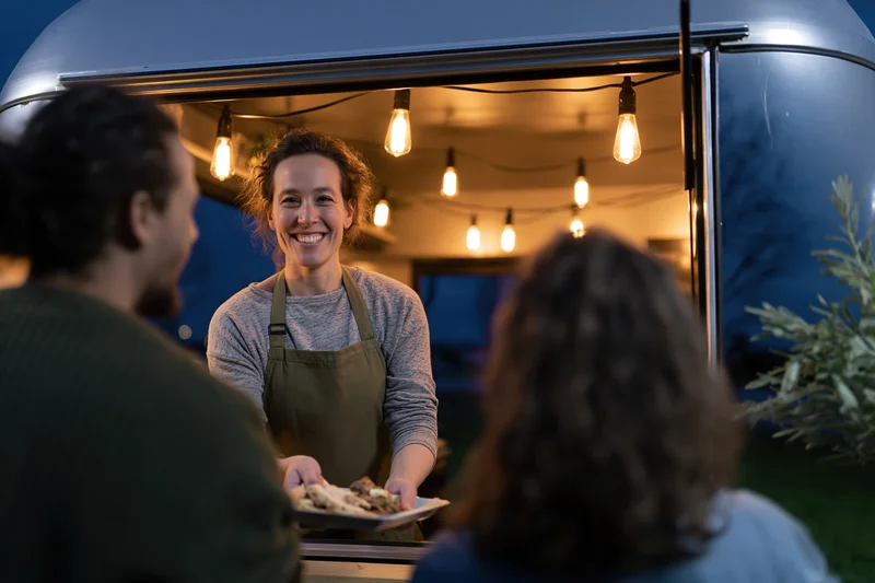 Woman in apron smiling and serving food from a food truck at night to customers outside