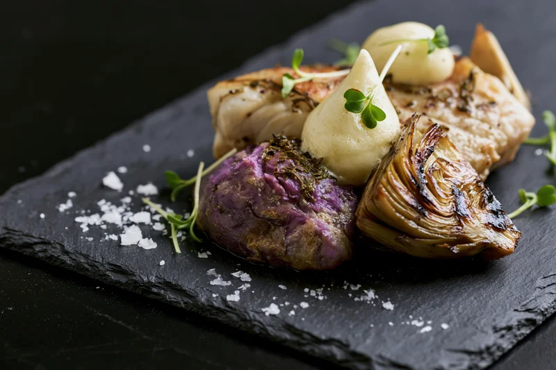 Roasted fish and vegetables artfully arranged on a dark slate board with microgreens and flaky salt