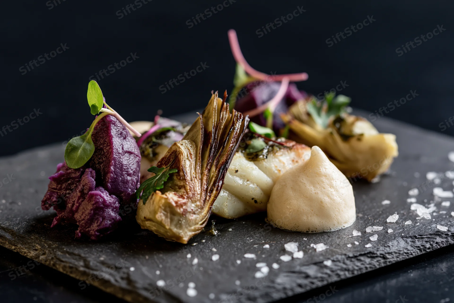 Gourmet dish with roasted artichoke, scallop, purple potato and sauce on dark slate board