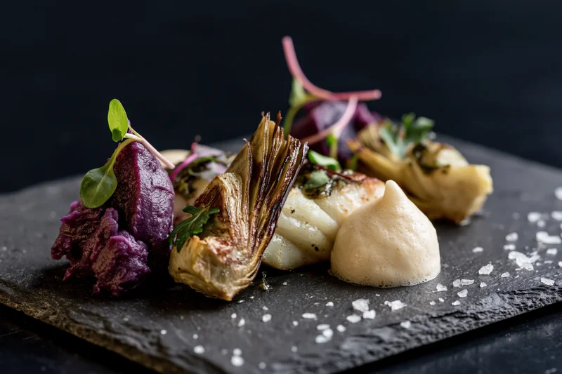 Gourmet dish with roasted artichoke, scallop, purple potato and sauce on dark slate board