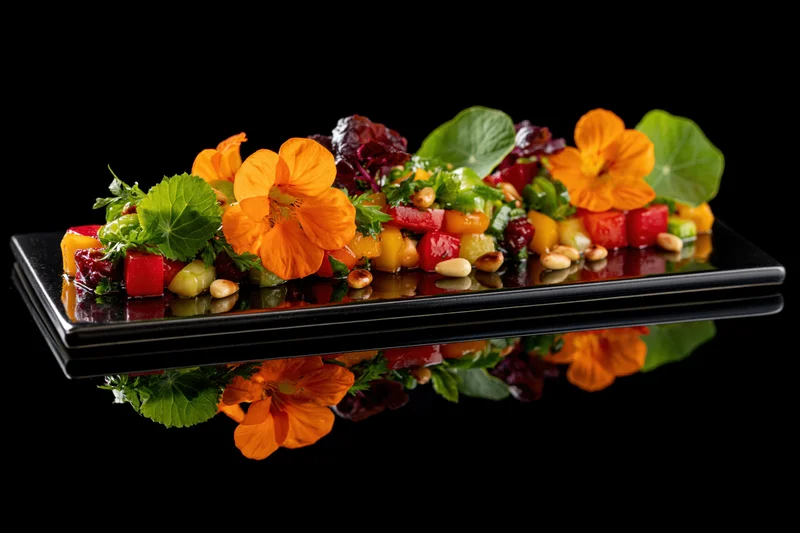 Colorful vegetable salad with edible orange nasturtium flowers and green leaves arranged on a reflective black plate