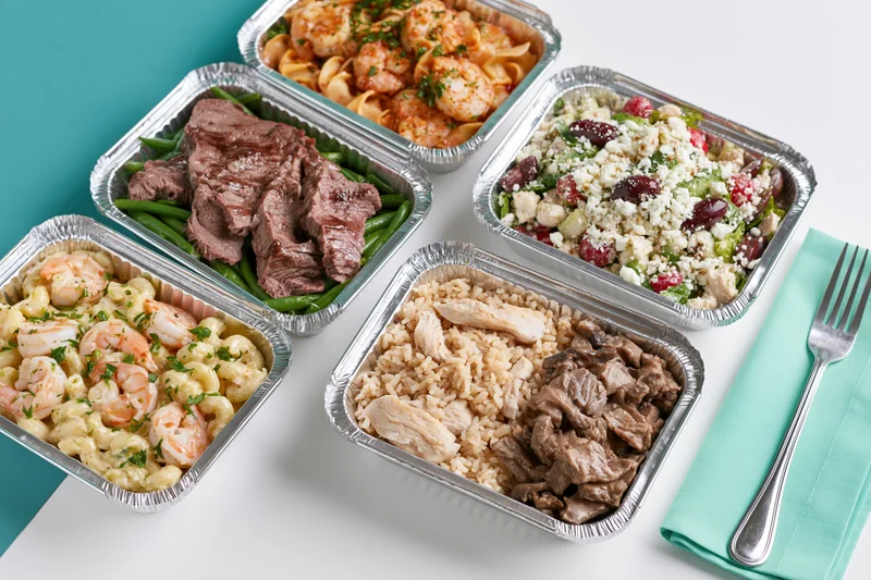 Five aluminum foil containers with prepacked meals arranged on a white surface with a teal napkin and fork beside them