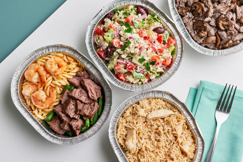 Four aluminum foil takeout containers with assorted meals arranged on a white surface with a teal napkin and fork