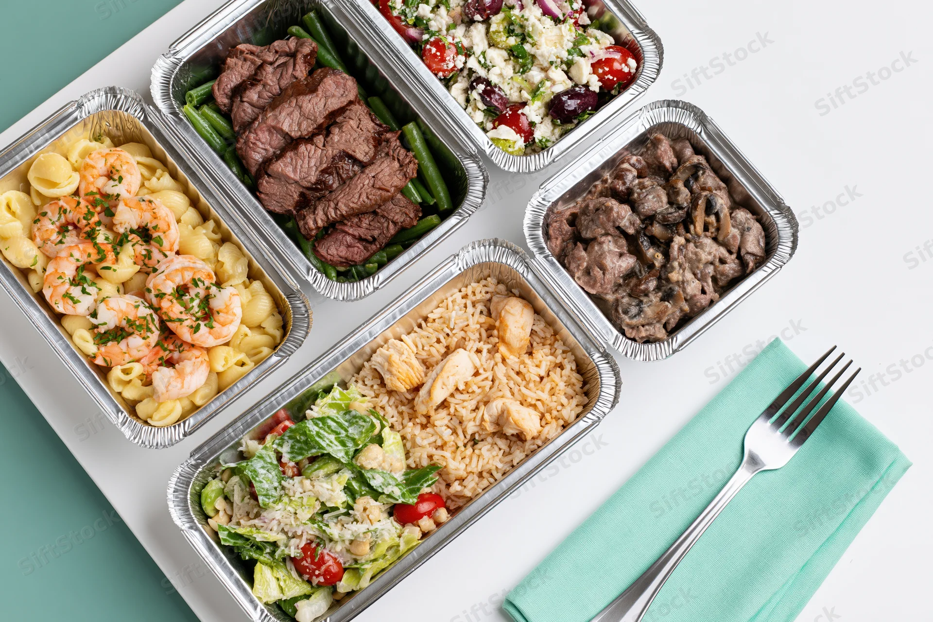 Five aluminum foil containers with prepacked meals arranged on a white surface with a fork and teal napkin