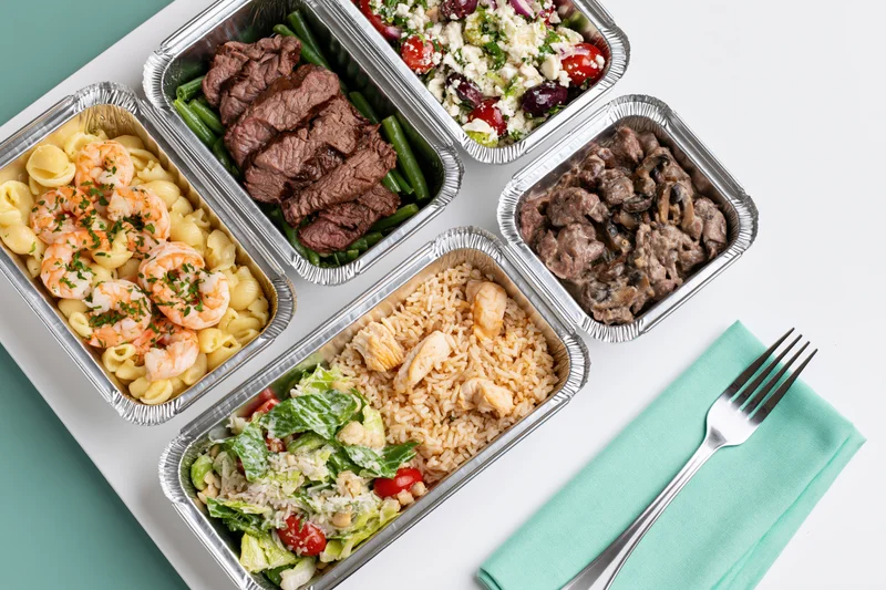 Five aluminum foil containers with prepacked meals arranged on a white surface with a fork and teal napkin