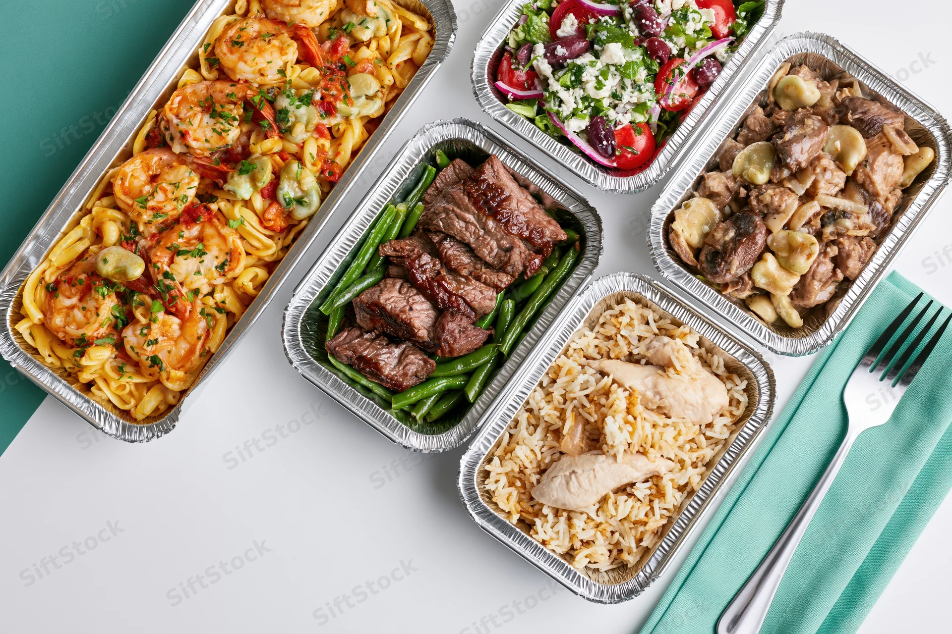 Five aluminum foil takeout containers with different meals arranged on a white and teal surface with a fork and napkin nearby