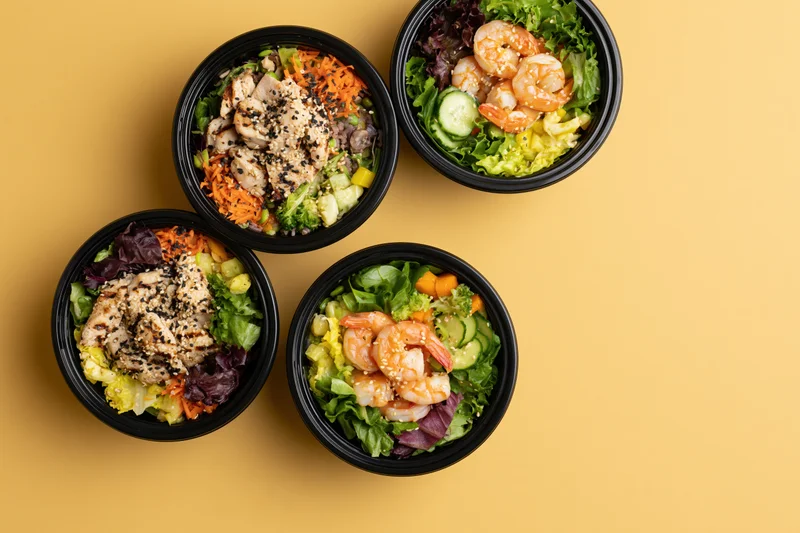Four black takeout bowls filled with poke salads featuring shrimp and chicken on a yellow background