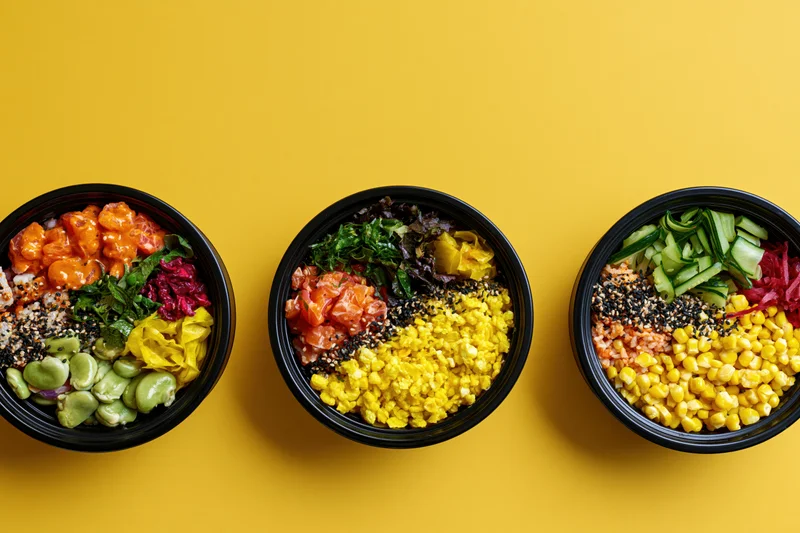 Three black bowls filled with colorful poke and grain salads arranged in a row on a yellow surface