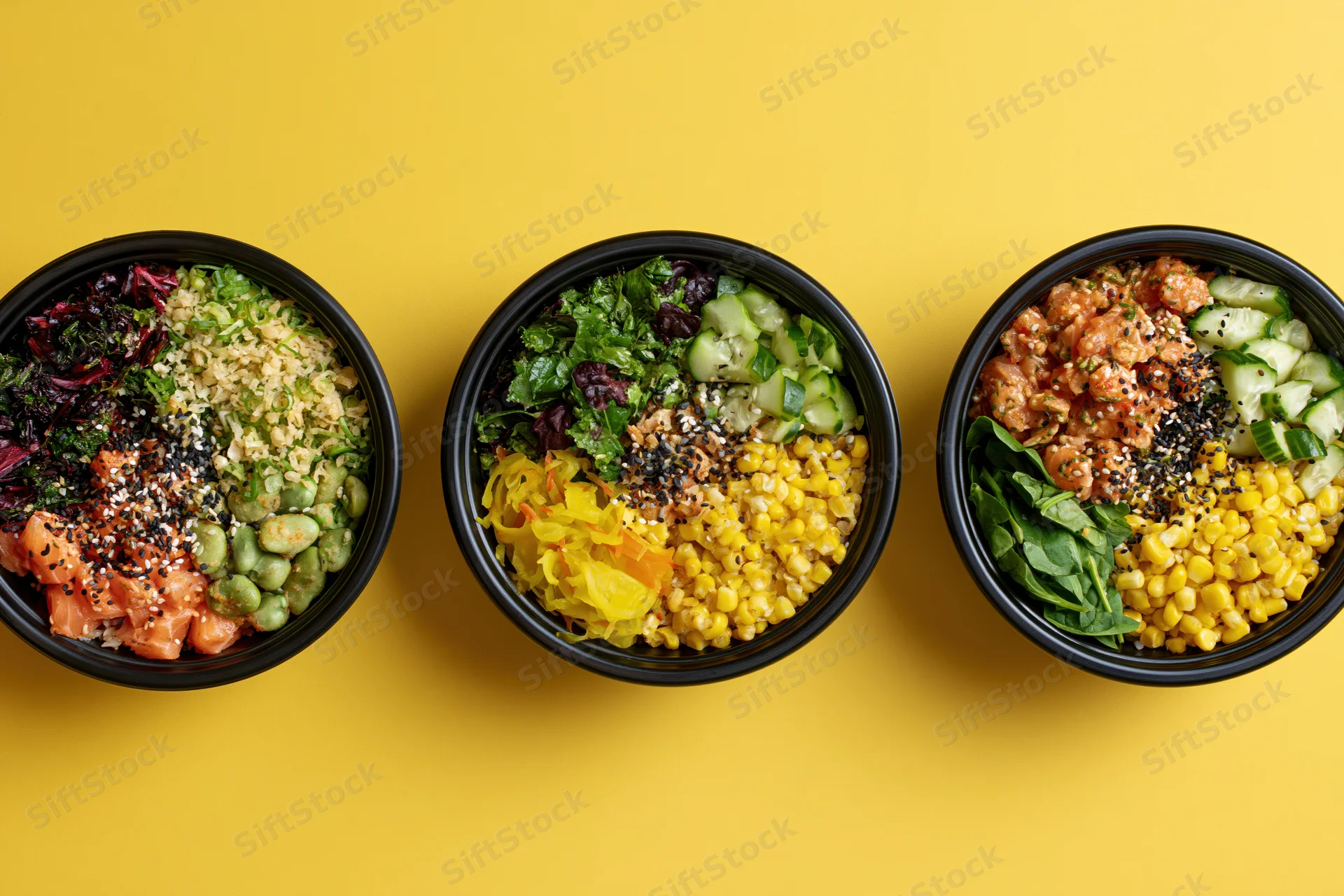 Three black bowls filled with poke and grain salads arranged in a row on a yellow surface