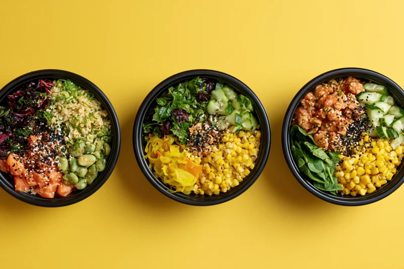 Three black bowls filled with poke and grain salads arranged in a row on a yellow surface