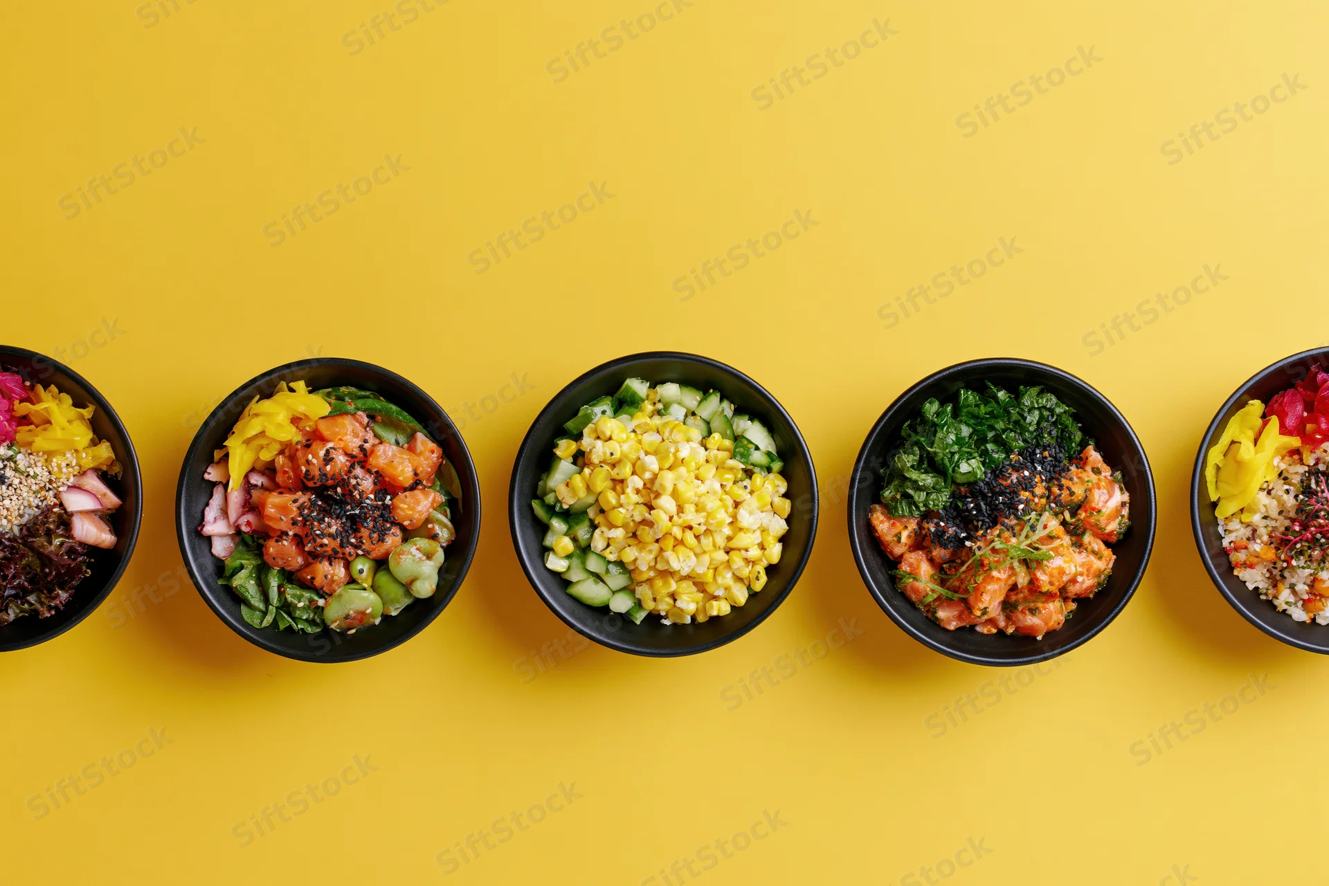 Five black bowls filled with poke and grain salads arranged in a row on a yellow surface