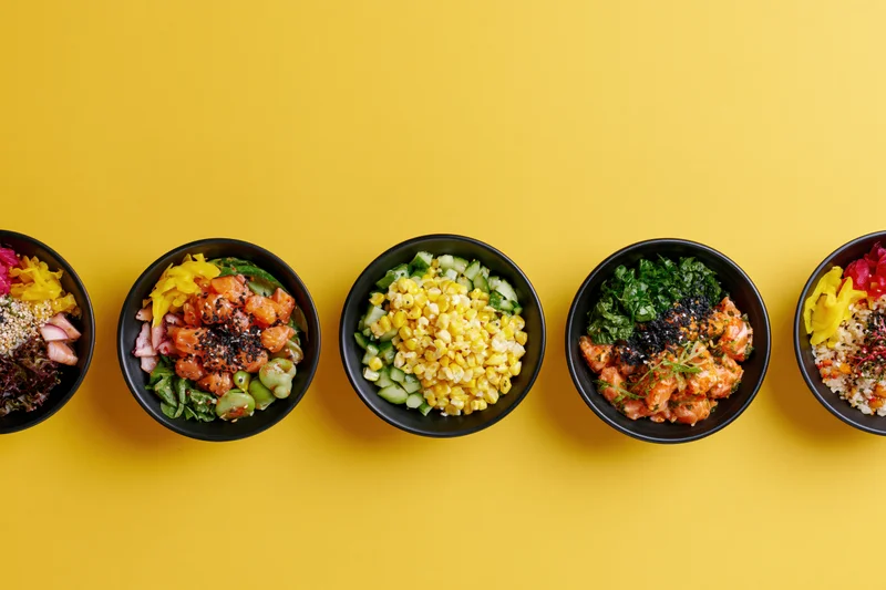 Five black bowls filled with poke and grain salads arranged in a row on a yellow surface