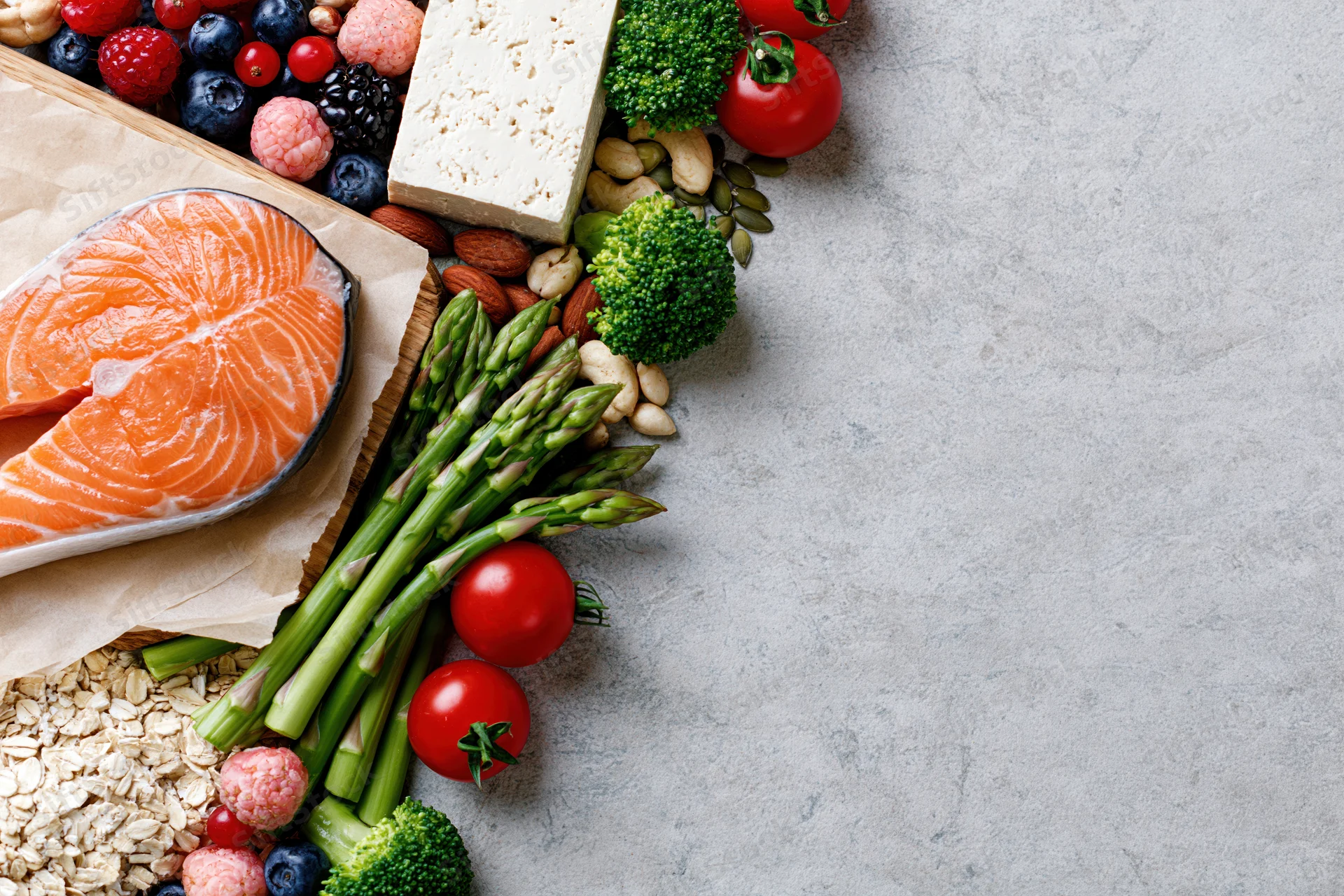 Fresh salmon, asparagus, broccoli, berries, cheese, nuts and oats arranged along the right edge of a gray surface