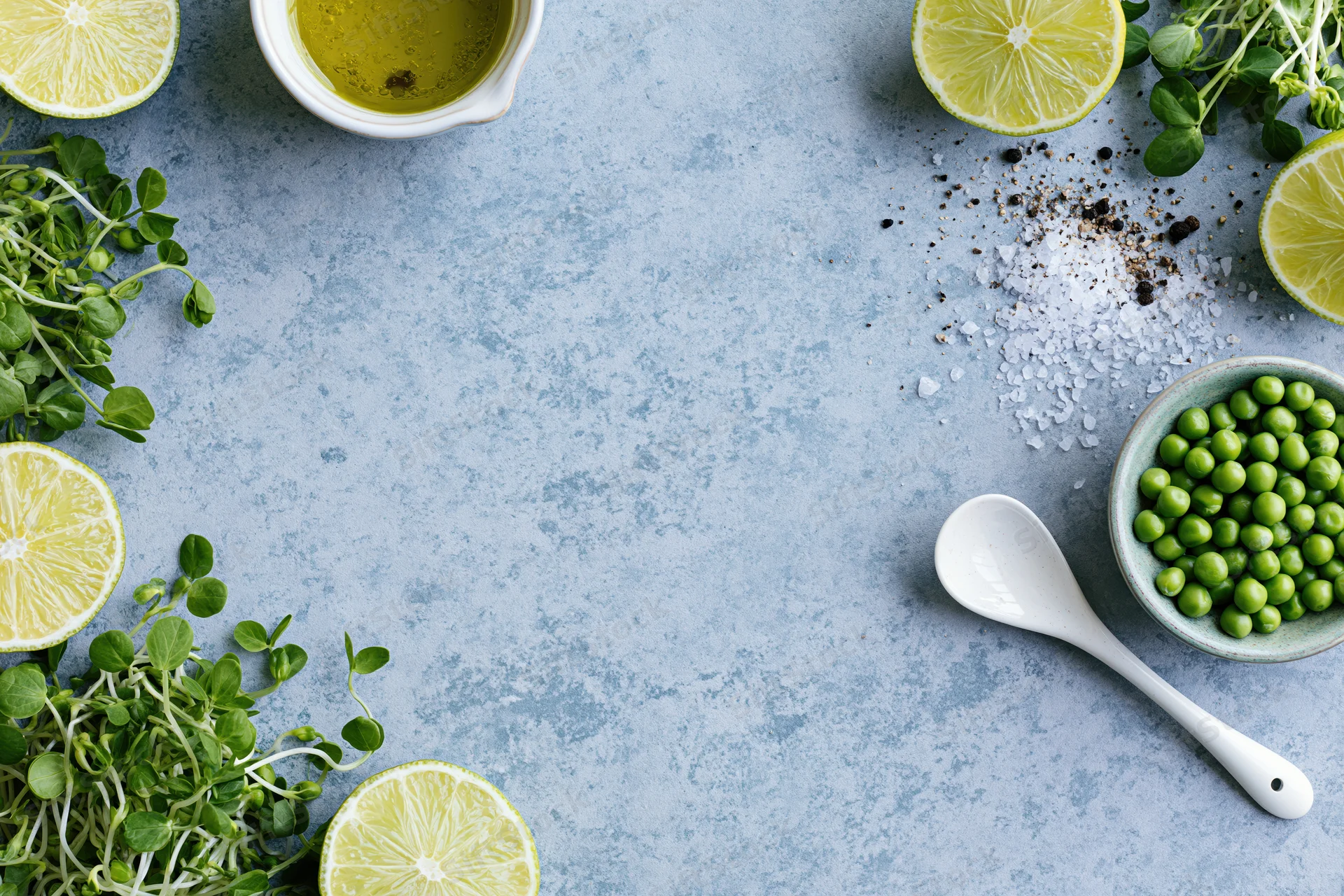 Flat lay arrangement of limes, peas, microgreens, salt, pepper, and a spoon on a pale blue-grey concrete surface