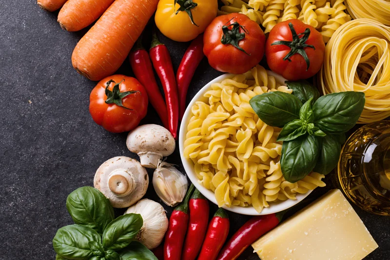 Fusilli pasta in a white bowl surrounded by fresh vegetables herbs cheese and olive oil on dark surface