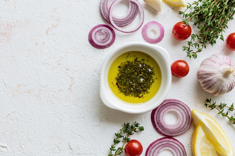 Small white ceramic bowl filled with olive oil and herbs surrounded by garlic, red onion, cherry tomatoes, lemon, and thyme on textured white surface