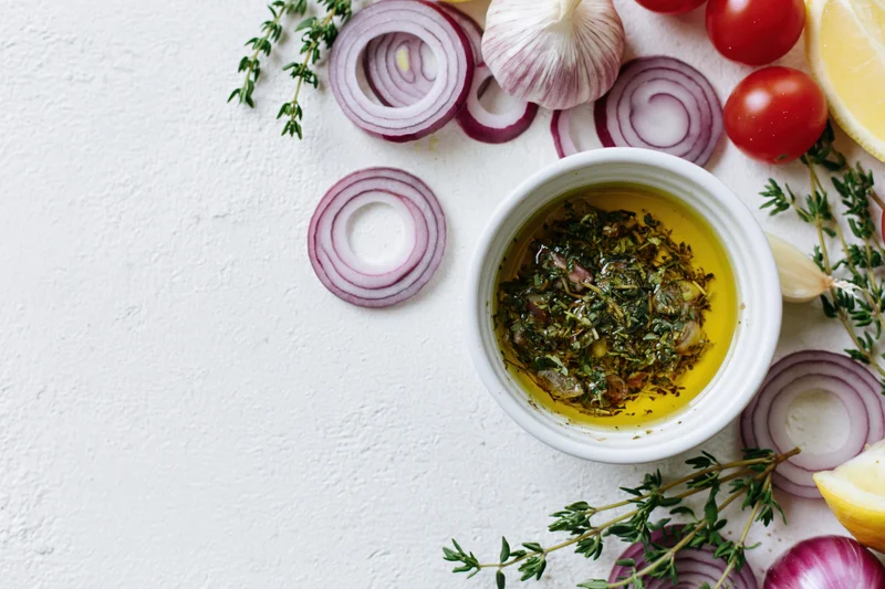 Small white ceramic bowl filled with olive oil and herbs surrounded by garlic, red onion, tomatoes, lemon, and thyme on a white surface