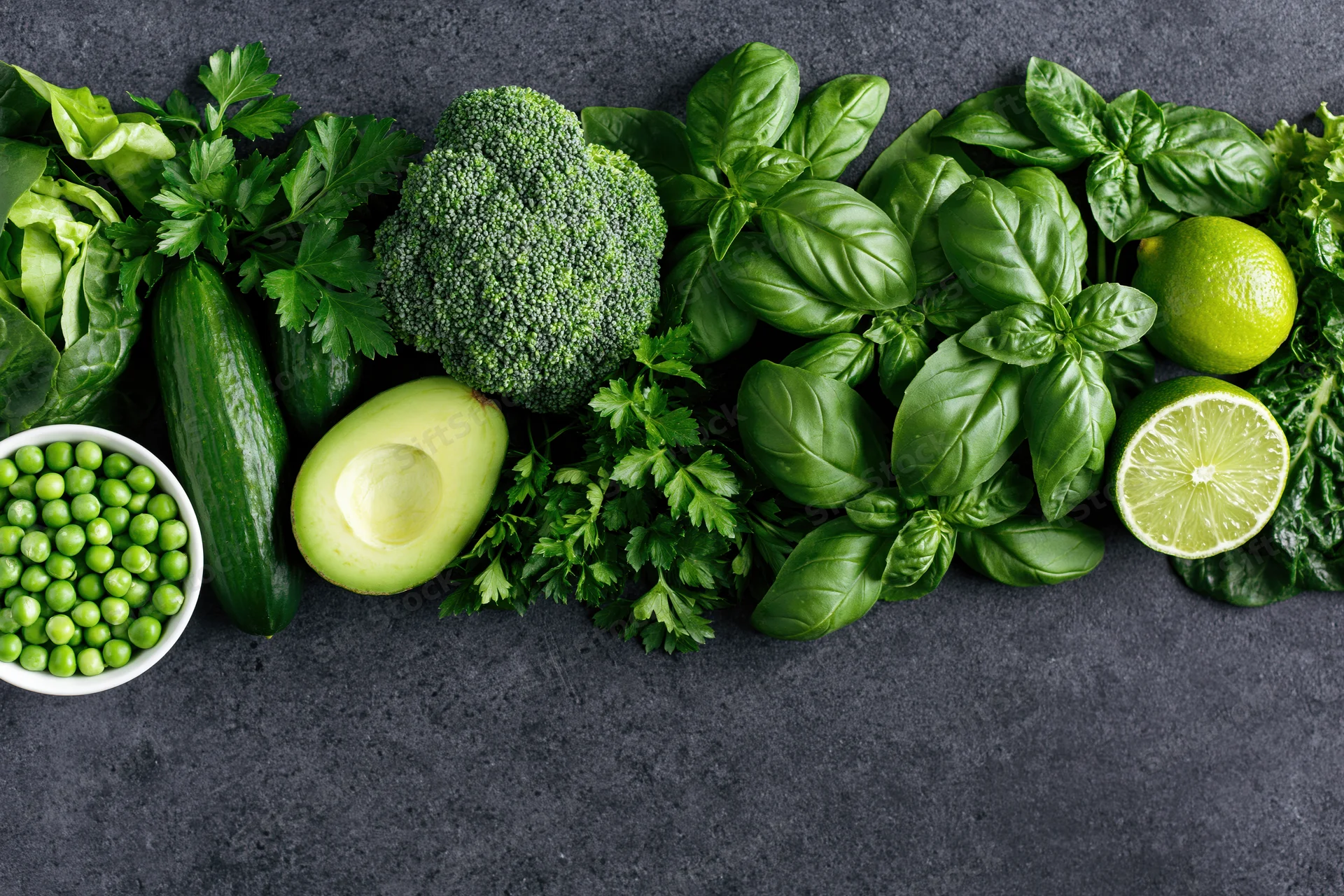 Assorted green vegetables and herbs arranged in a flat lay on dark textured surface