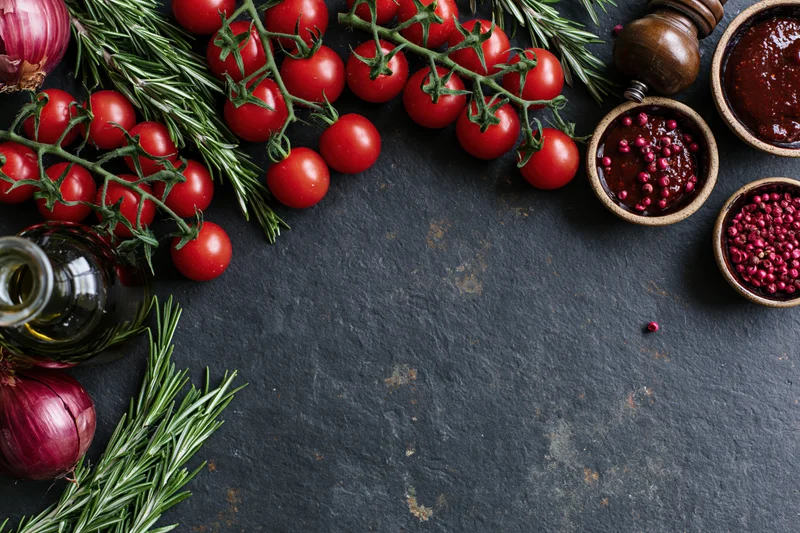 Cherry tomatoes on the vine with rosemary, red onion, olive oil and spices arranged on a dark slate surface
