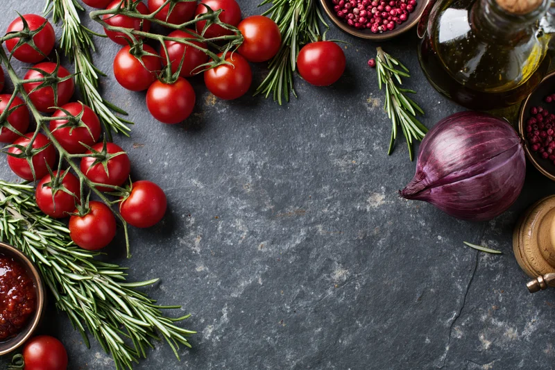 Cherry tomatoes on the vine with rosemary, red onion, olive oil and peppercorns arranged on slate surface