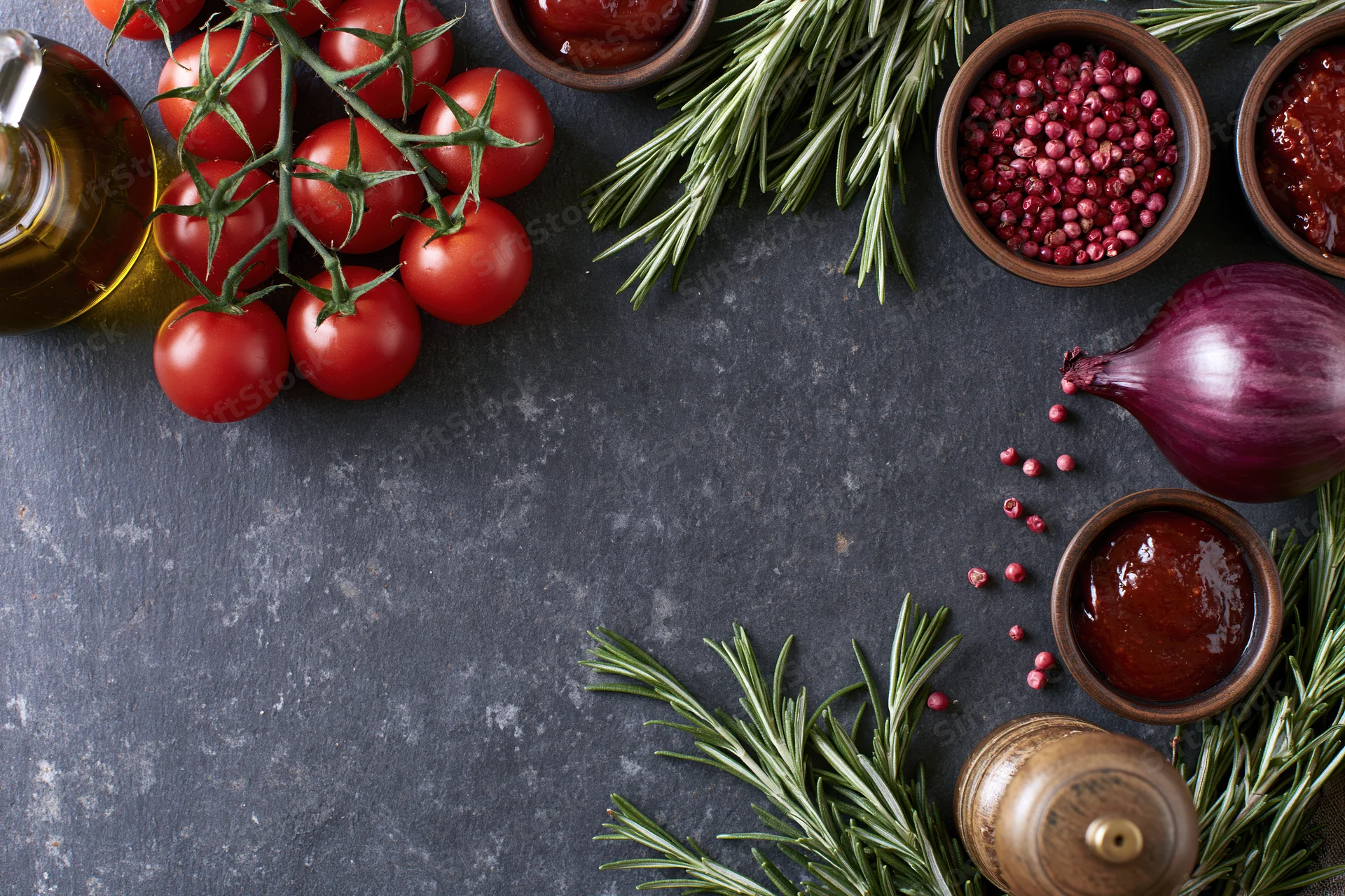 Cherry tomatoes on the vine with rosemary sprigs, red onion, spices and olive oil arranged on dark slate surface