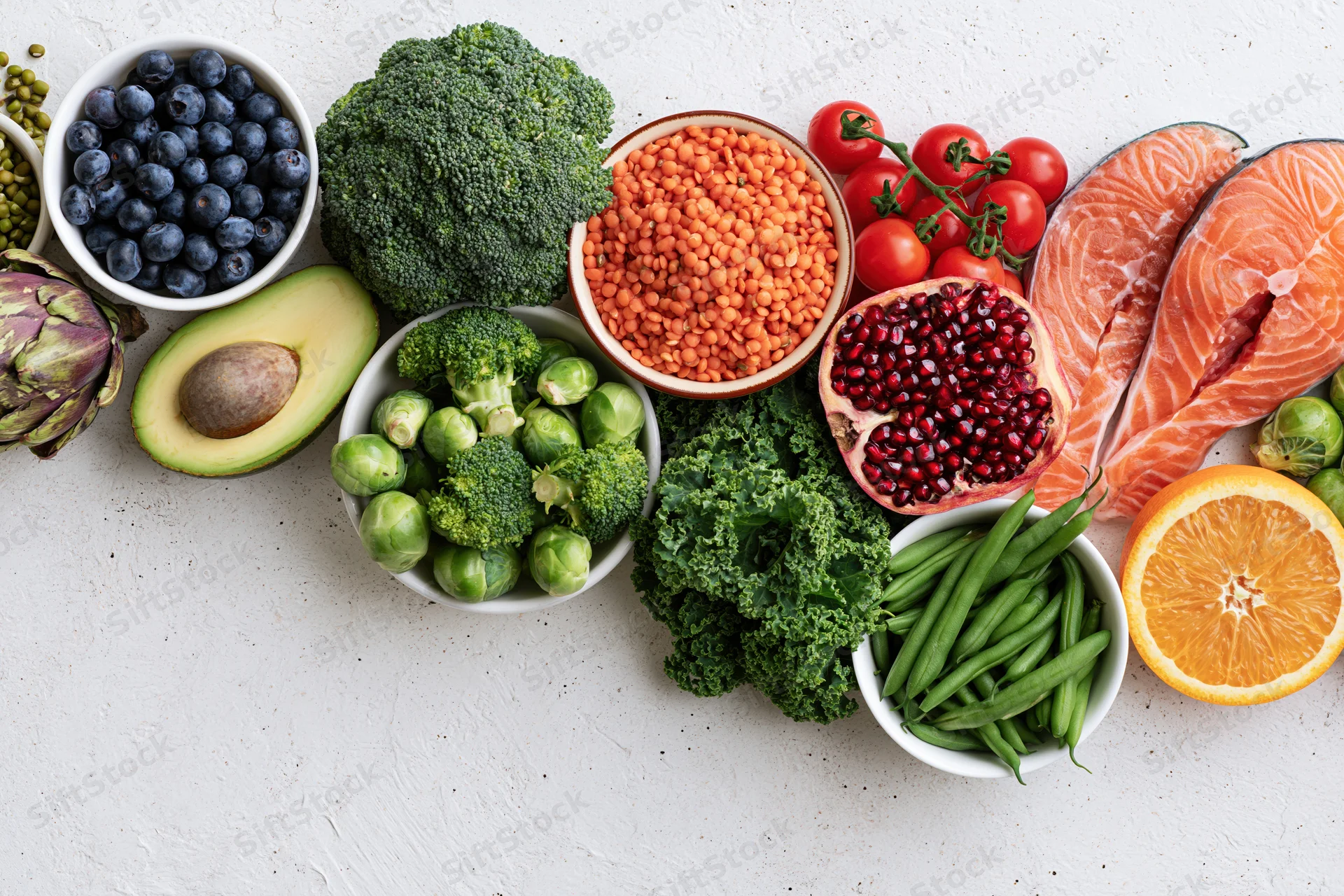 Fresh healthy foods including salmon, broccoli, blueberries, avocado, lentils, tomatoes, pomegranate, kale, green beans, orange, brussels sprouts, and artichoke arranged in a flat lay on a white textured surface