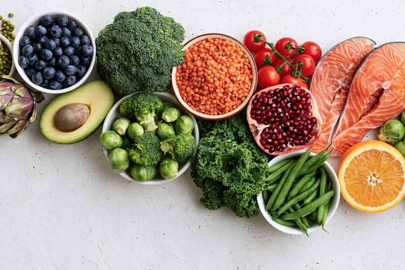 Fresh healthy foods including salmon, broccoli, blueberries, avocado, lentils, tomatoes, pomegranate, kale, green beans, orange, brussels sprouts, and artichoke arranged in a flat lay on a white textured surface
