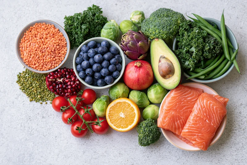Fresh healthy foods including salmon, vegetables and fruits arranged in a flat lay on a white textured surface
