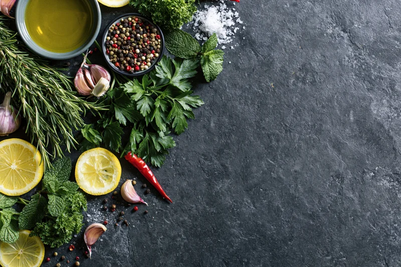 Fresh herbs and spices arranged along the left side of a dark stone surface with copyspace on the right