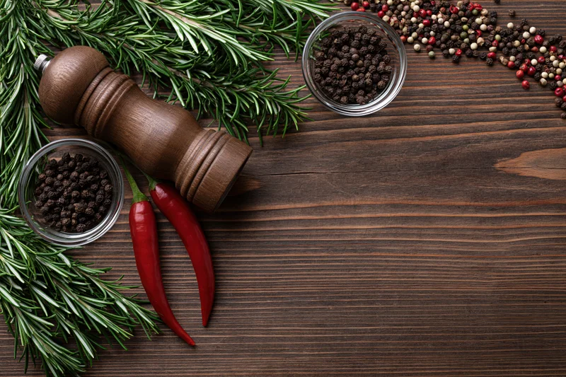 Fresh rosemary sprigs, red chili peppers, peppercorns, and a wooden grinder arranged on a dark wooden table in a flat lay composition