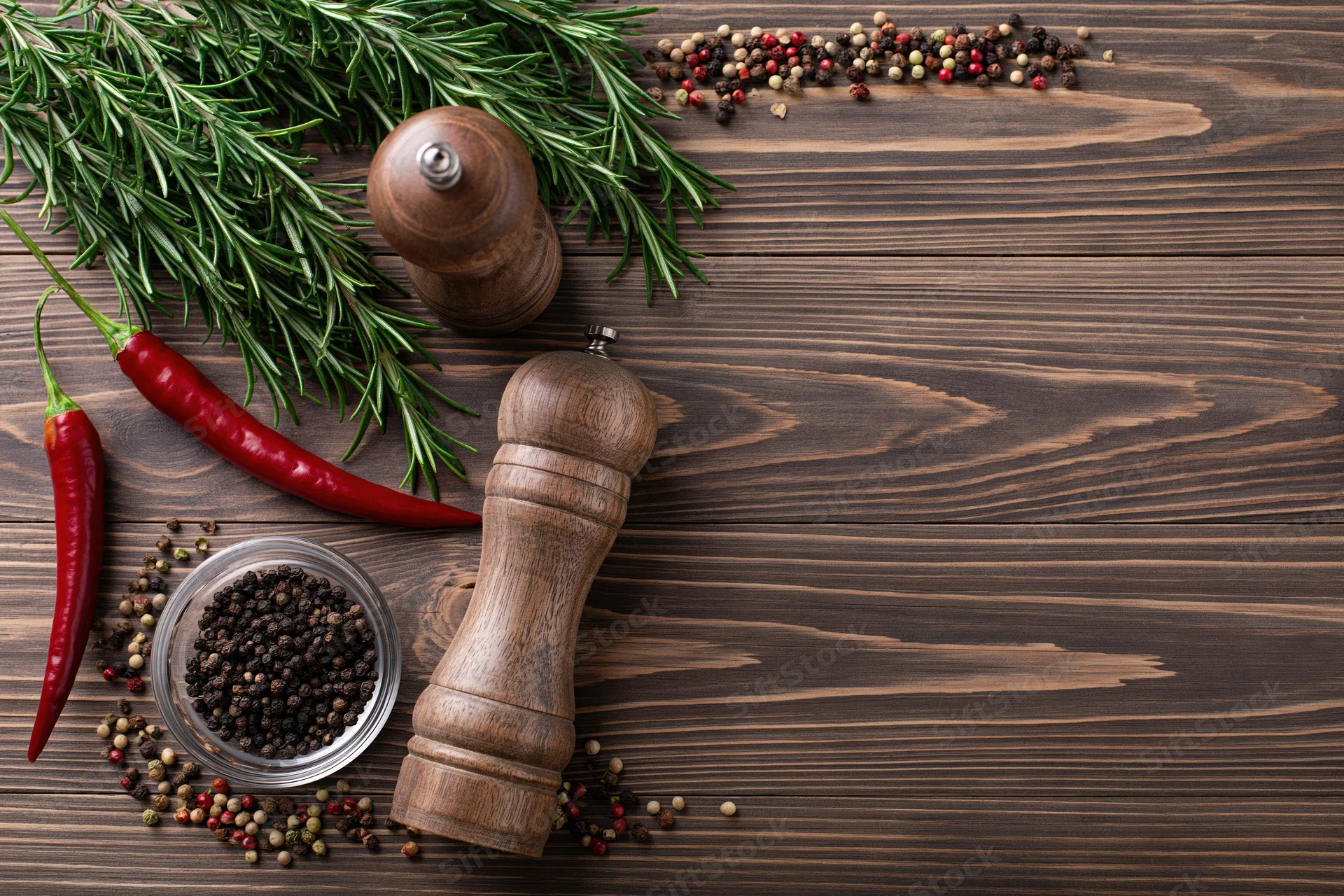 Fresh rosemary sprigs, red chili peppers, and wooden pepper grinders arranged on a dark wooden table with scattered peppercorns and a small bowl, viewed from above