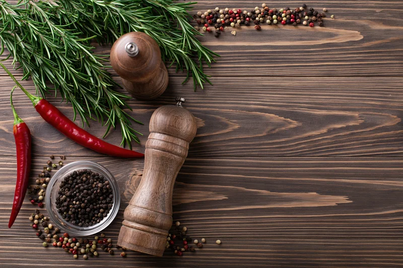 Fresh rosemary sprigs, red chili peppers, and wooden pepper grinders arranged on a dark wooden table with scattered peppercorns and a small bowl, viewed from above
